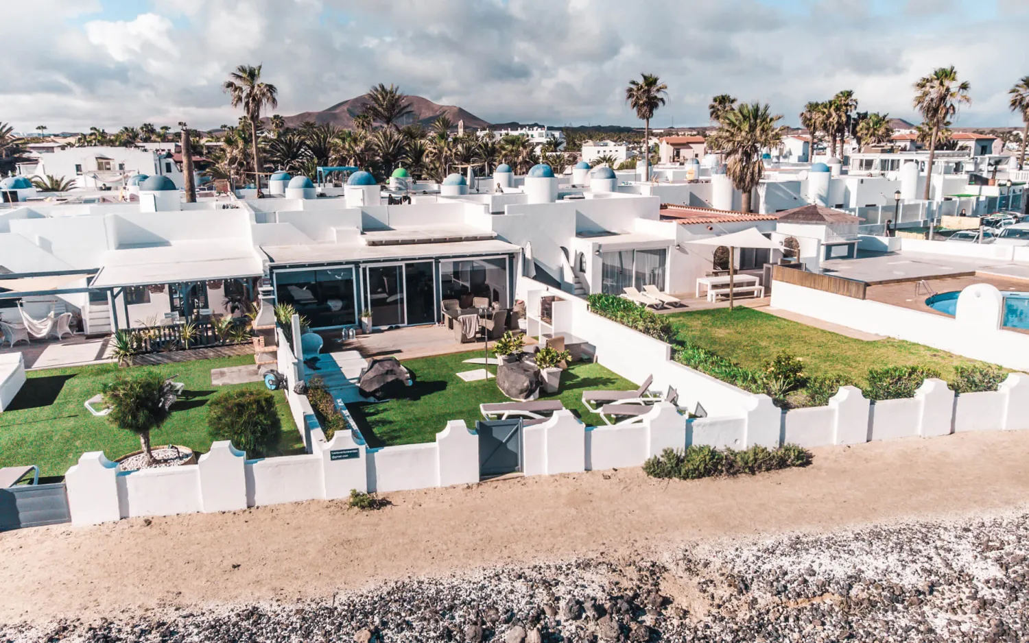Luftaufnahme einer weißen Ferienanlage an der Küste von Fuerteventura mit Rasenflächen, Palmen und Pool, im Hintergrund ein Vulkanberg